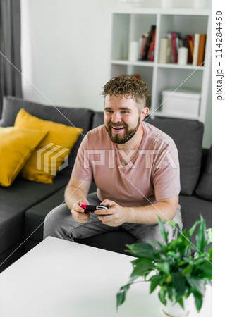 Cheerful young man seated on a sofa playing video games at home. Gaming addiction and entertainment 114284450
