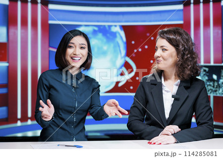 Morning show hosts debate events, presenting latest news on international television network in newsroom. Tewo colleagues working on entertainment segment newscast doing reportage. 114285081