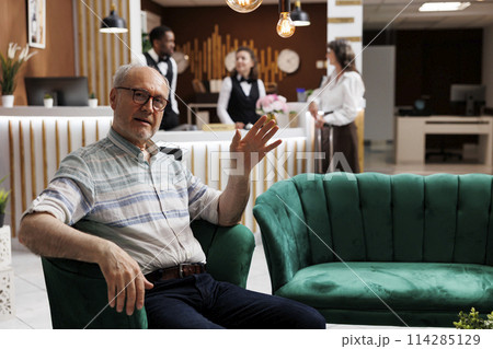 Portrait shot of old man seated on cozy sofa in exclusive hotel reception, waving at camera. Comfortable lobby offers relaxing lounge area for elderly male traveler waiting for check-in procedure. Portrait shot of old man seated on cozy sofa in exclusive hotel reception, waving at camera. Comfortable lobby offers relaxing lounge area for elderly male traveler waiting for check-in procedure. 114285129