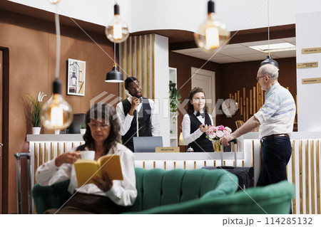 With assistance from caucasian receptionist in hotel lobby, retired senior man with suitcase checks in at front desk. While male visitor waits at counter, an elderly woman reading in cozy lounge area. 114285132