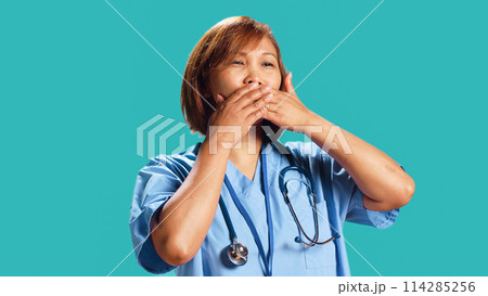 Certified nurse covering mouth with hands, holding back from saying rude thoughts. Hospital employee silencing herself, abstaining from speaking, isolated over studio background, close up 114285256