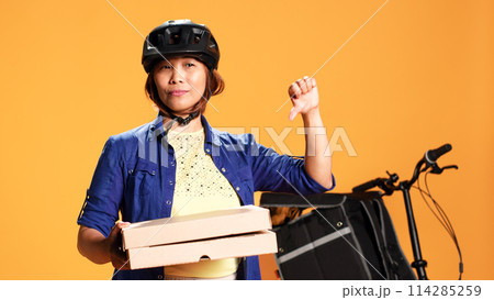 Close up shot of part time courier holding takeaway pizza boxes, showing thumbs down sign. Food delivery cyclist doing disapproving finger shaking gesture, saying no, isolated over studio background Close up shot of part time courier holding takeaway pizza boxes, showing thumbs down sign. Food delivery cyclist doing disapproving finger shaking gesture, saying no, isolated over studio background 114285259