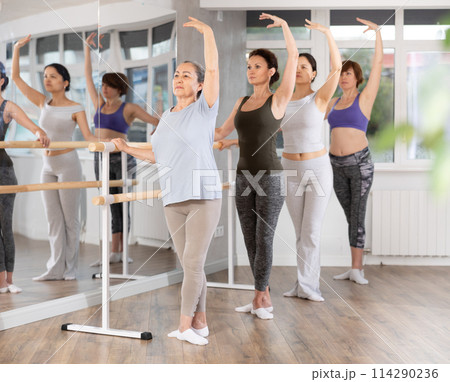Female amateurs stand in third position near ballet barre during group training in dance studio Female amateurs stand in third position near ballet barre during group training in dance studio 114290236