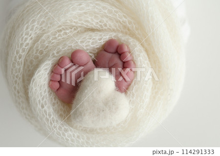 The tiny foot of a newborn baby. Soft feet of a new born in a white wool blanket. Close up of toes, heels and feet of a newborn. Knitted white heart in the legs of a baby. Macro photography 114291333