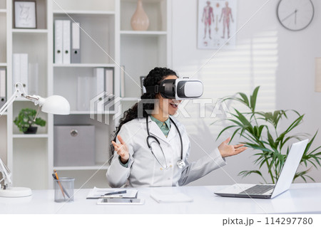 Excited Hispanic female doctor using virtual reality headset at her desk in a well-equipped medical office, exploring new technology in healthcare. Excited Hispanic female doctor using virtual reality headset at her desk in a well-equipped medical office, exploring new technology in healthcare. 114297780