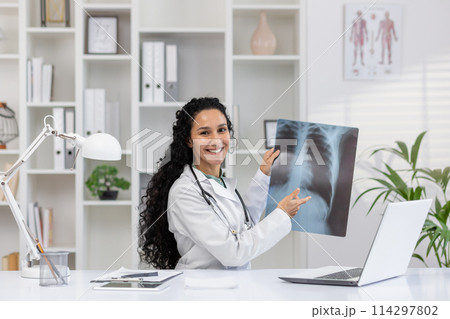A professional female radiologist in a white lab coat is joyfully examining and explaining a chest X-ray in her well-equipped clinic office, surrounded by medical charts and a laptop. A professional female radiologist in a white lab coat is joyfully examining and explaining a chest X-ray in her well-equipped clinic office, surrounded by medical charts and a laptop. 114297802