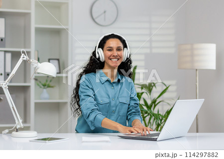 A cheerful Hispanic woman wearing headphones works at her desk in a bright modern office, interacting with her laptop, embodying professionalism and happiness. 114297882