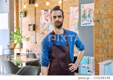 Young man worker, owner in apron looking at camera in restaurant, coffee shop Young man worker, owner in apron looking at camera in restaurant, coffee shop 114298146