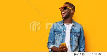 Happy African millennial man laughing with mobile phone on yellow studio backdrop. Joyful black man wearing jeans jacket, sunglasses smiling looking aside. Great new mobile App. People and gadgets 114304859