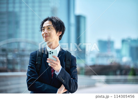 スマホを操作するリュックを担いだスーツ姿のビジネスマン スマホを操作するリュックを担いだスーツ姿のビジネスマン 114305456