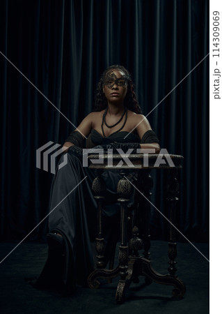 Confident, statuesque African-American woman seated in classic vintage black dress with mask against dark curtain backdrop. 114309069