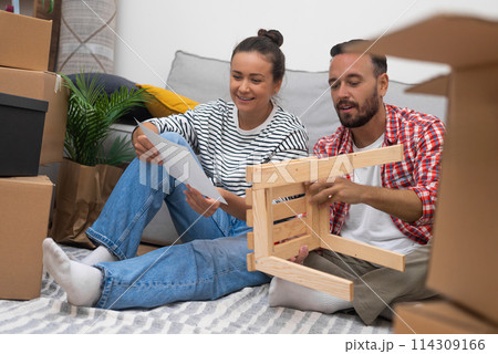 Happy newlyweds install furniture in living room sitting on floor among collected decorations on territory of modern apartment received as wedding gift from parents Happy newlyweds install furniture in living room sitting on floor among collected decorations on territory of modern apartment received as wedding gift from parents 114309166