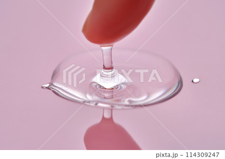 A finger touches a drop of cosmetic product on a pink background. A finger touches a drop of cosmetic product on a pink background. 114309247