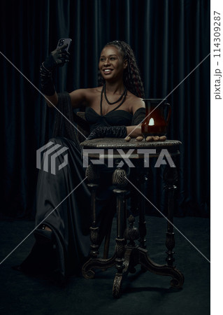 Young African-American woman sitting on chair in vintage style dress taking selfie using modern smartphone against dark curtain backdrop. Young African-American woman sitting on chair in vintage style dress taking selfie using modern smartphone against dark curtain backdrop. 114309287