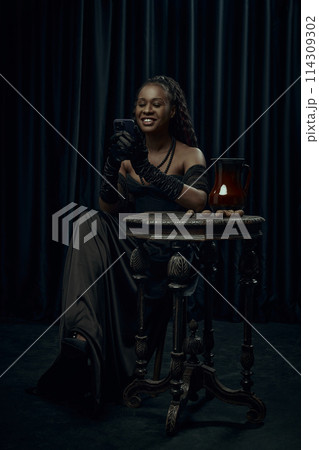 Young African-American woman sitting on chair in vintage style dress and using modern smartphone against dark curtain backdrop. Young African-American woman sitting on chair in vintage style dress and using modern smartphone against dark curtain backdrop. 114309302