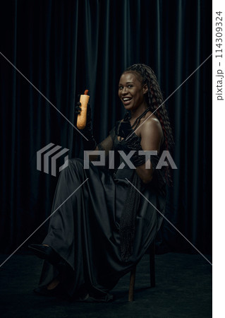 Shocked young African-American woman dressed as medieval person holds fast food, hot-dog and shows thumb up against dark curtain backdrop. 114309324