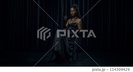 African-American woman, dressed old-fashion dress, looks medieval person, sits confidently on chair holding glass of brandy against dark curtain backdrop. 114309429