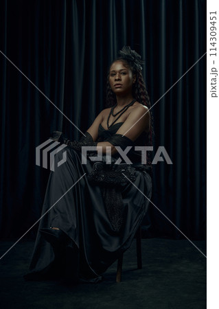 African-American woman, dressed old-fashion dress, looks as princess, sits confidently on chair against dark curtain backdrop. 114309451