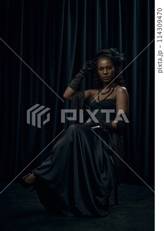 Regal young woman with long, braided hair sits confidently on chair, wearing old-fashion flowing black gown against dark curtain backdrop. Regal young woman with long, braided hair sits confidently on chair, wearing old-fashion flowing black gown against dark curtain backdrop. 114309470