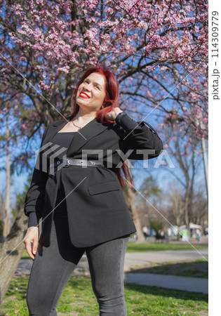 Fashion outdoor photo of beautiful woman with red curly hair in elegant suit posing in spring flowering park with blooming cherry tree. Copy space and empty place for advertising text 114309779