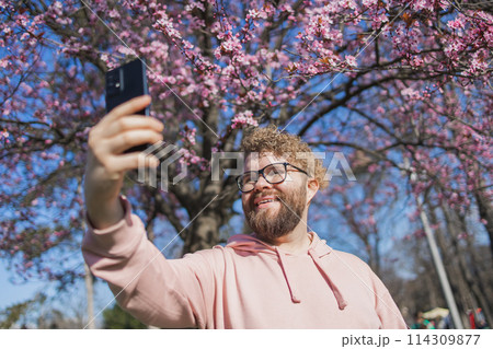 Happy curly man takes selfie against backdrop of flowering tree in spring for his internet communications. Weekend and social networks concept 114309877