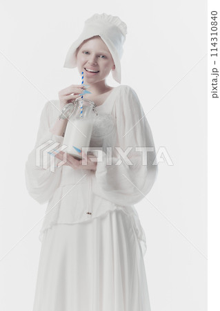 Smiling young woman dressed as medieval person, maid in white historic dress drinks milk against white studio background. 114310840