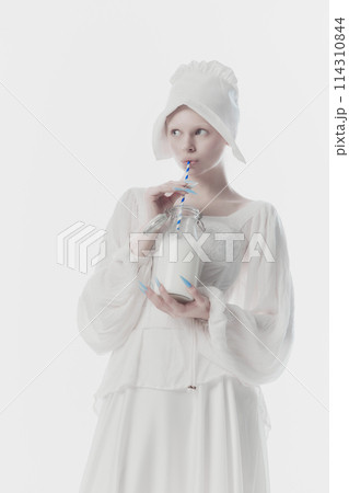 Beautiful young woman dressed as medieval person, maid in white historic dress drinks milk against white studio background. Beautiful young woman dressed as medieval person, maid in white historic dress drinks milk against white studio background. 114310844