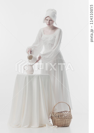 Young woman dressed as medieval person, maid in white dress poring milk into jug against white studio background. 114310855