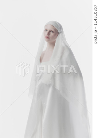 Young, calm woman shrouded in gossamer white fabric, creating ghostly yet angelic appearance against white studio background. 114310857
