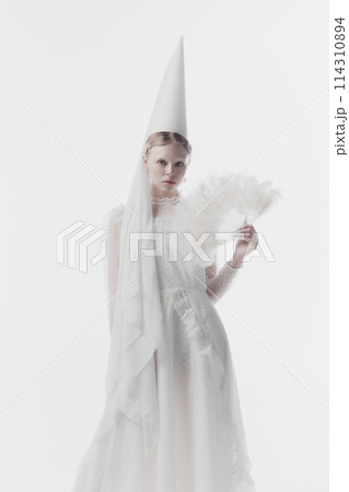 Portrait of young woman in white costume with conical hat, looks as medieval person, holding feathery fan against white studio background. Portrait of young woman in white costume with conical hat, looks as medieval person, holding feathery fan against white studio background. 114310894