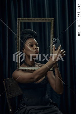 African-American woman sitting posing behind ornate, vintage frame and taking selfie using modern smartphone against dark curtain backdrop. 114310947