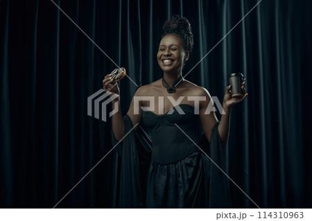 Smiling African-American woman, dressed in vintage black dress holds sweet donut and cup of coffee against dark curtain backdrop. Smiling African-American woman, dressed in vintage black dress holds sweet donut and cup of coffee against dark curtain backdrop. 114310963