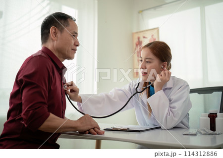 Female doctor asian cardiologist examining senior male cardiac patient listening checking heartbeat using stethoscope at checkup hospital visit. elderly healthcare cardiology concept 114312886