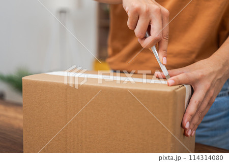 Woman buyer uses stationery knife to cut cardboard box with orders from online store with home delivery female hands open package in light accommodation 114314080