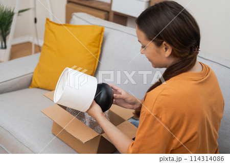 Woman takes out small table lamp from cardboard parcel with home delivery online purchase from furniture store and decorative element with lighting function 114314086