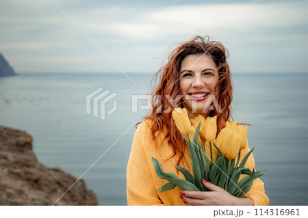 Portrait of a happy woman with hair flying in the wind against the backdrop of mountains and sea. Holding a bouquet of yellow tulips in her hands, wearing a yellow sweater Portrait of a happy woman with hair flying in the wind against the backdrop of mountains and sea. Holding a bouquet of yellow tulips in her hands, wearing a yellow sweater 114316961