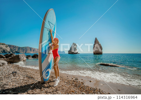 Woman surfer during summer travel to beach vacation. A middle-aged woman in a red swimsuit and a cap with sap on the background of the sea and rocks in the water. extreme sport. Travel, weekend Woman surfer during summer travel to beach vacation. A middle-aged woman in a red swimsuit and a cap with sap on the background of the sea and rocks in the water. extreme sport. Travel, weekend 114316964