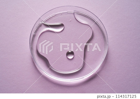 Serum or cosmetic oil flows into a transparent bowl on a purple background. 114317125