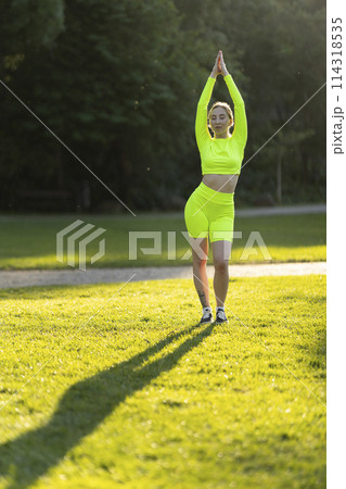 A woman in a neon green outfit is doing yoga on a grassy field 114318535
