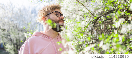 Banner Male bearded man standing under branches with flowers of blooming almond or cherry tree in spring garden. Spring blossom. Copy space Banner Male bearded man standing under branches with flowers of blooming almond or cherry tree in spring garden. Spring blossom. Copy space 114318950
