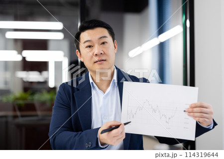 Professional Asian businessman in a suit displaying a financial report chart, engaging with the camera in a modern office setting. 114319644