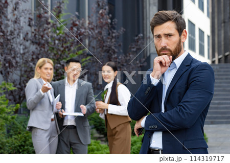 Portrait of upset young man in business suit standing outside office building looking disappointed at camera, behind group of interracial people cheering and laughing at new colleague. 114319717