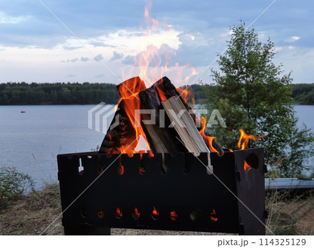 Orange flame of fire on firewood and coals in a barbecue on the shore of a lake with calm water in nature in the forest against the background of clouds in the evening in summer on vacation 114325129