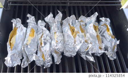 Several ears of corn wrapped in aluminum foil are lined up on a barbecue grill, slowly roasting to perfection, showcasing a popular and delicious method of cooking this classic side dish. Several ears of corn wrapped in aluminum foil are lined up on a barbecue grill, slowly roasting to perfection, showcasing a popular and delicious method of cooking this classic side dish. 114325854