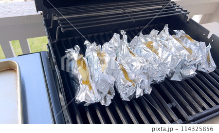 Several ears of corn wrapped in aluminum foil are lined up on a barbecue grill, slowly roasting to perfection, showcasing a popular and delicious method of cooking this classic side dish. 114325856