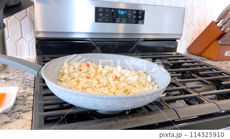 A frying pan on a gas stove sizzles with diced potatoes and colorful bits of red and green peppers, showcasing a delicious and simple home-cooked side dish in the making. 114325910