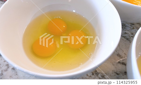 This image showcases three raw eggs cracked into a white bowl, set on a marble countertop, ready for mixing or cooking, highlighting the simplicity and beauty of basic cooking ingredients. 114325955