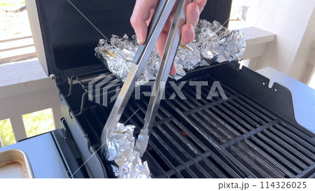 Several ears of corn wrapped in aluminum foil are lined up on a barbecue grill, slowly roasting to perfection, showcasing a popular and delicious method of cooking this classic side dish. 114326025