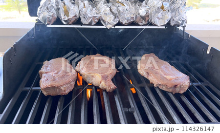 A person uses tongs to flip juicy steaks on a grill, with flames licking the meat, captured in a moment of intense heat and sizzle, showcasing a classic outdoor cooking experience. A person uses tongs to flip juicy steaks on a grill, with flames licking the meat, captured in a moment of intense heat and sizzle, showcasing a classic outdoor cooking experience. 114326147