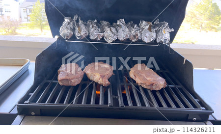 A person uses tongs to flip juicy steaks on a grill, with flames licking the meat, captured in a moment of intense heat and sizzle, showcasing a classic outdoor cooking experience. A person uses tongs to flip juicy steaks on a grill, with flames licking the meat, captured in a moment of intense heat and sizzle, showcasing a classic outdoor cooking experience. 114326211